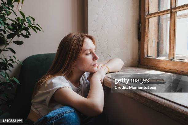 upset redhead teen girl sitting by window looking at phone waiting call or message - unhappy stockfoto's en -beelden