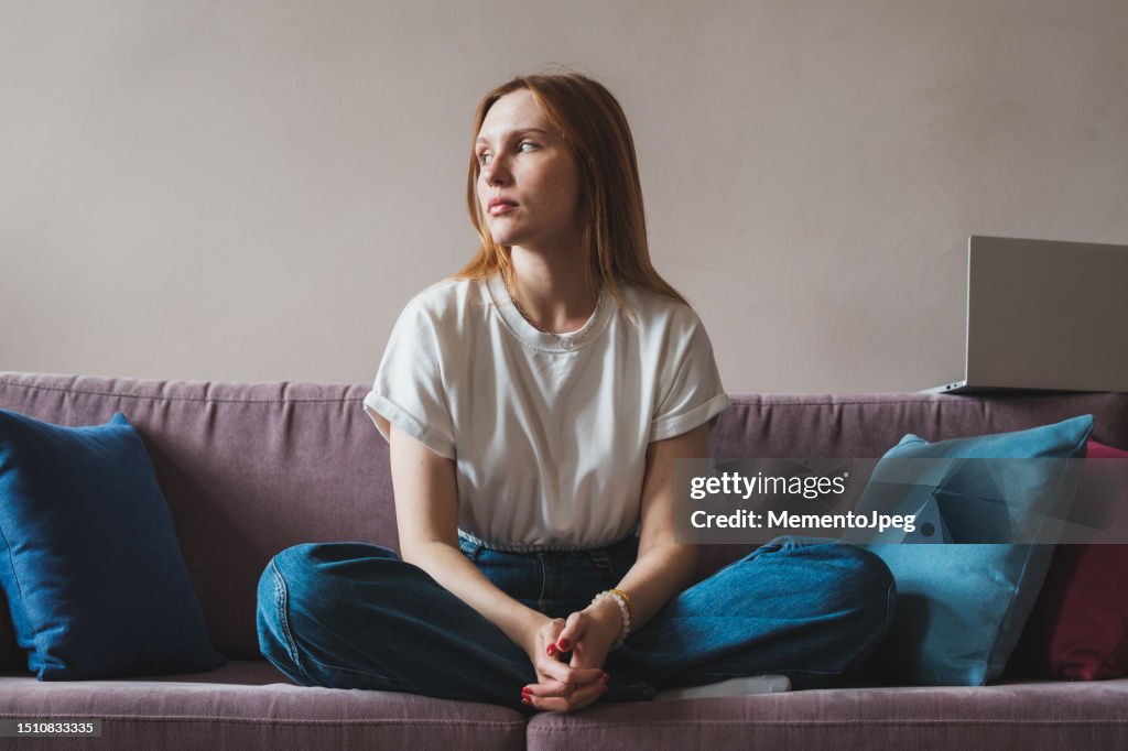 Upset depressed young woman freelancer sitting on sofa with laptop looking out window