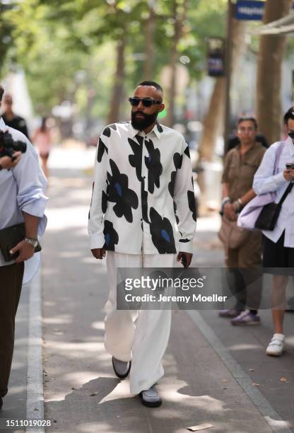 Jean-Claude Mpassy is seen wearing black sunglasses, a buttoned long-sleeved shirt in white with big flowers in black and blue on it, wide pants in...
