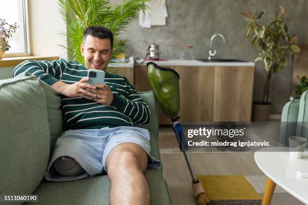young disabled man using a smart phone in the living room at home - amputee stock pictures, royalty-free photos & images