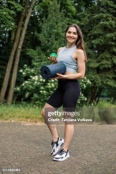 portrait of charismatic young woman in sports clothes and sneakers standing in park holding yoga roll and massage ball - massagebal stockfoto's en -beelden