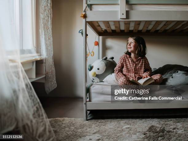 a little girl sits on a bottom bunk of a bed in a bedroom, holding a book and gazing towards the window - annual event stock pictures, royalty-free photos & images