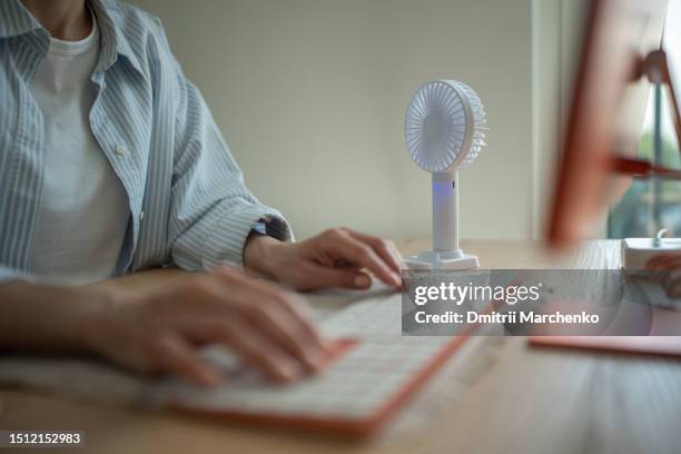 hands of woman typing on computer keyboard, portable compact fan cooling air near on desk. - hittegolf stockfoto's en -beelden