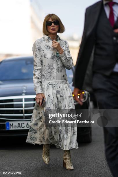 Anna Wintour wears black sunglasses, a white with black flower print pattern silk long sleeves / long dress, beige and brown shiny leather snake...