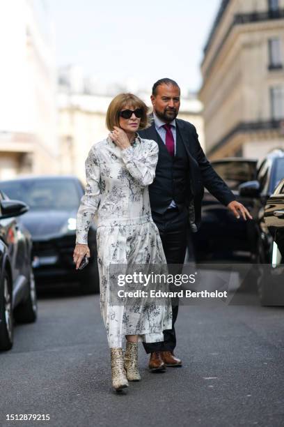 Anna Wintour wears black sunglasses, a white with black flower print pattern silk long sleeves / long dress, beige and brown shiny leather snake...