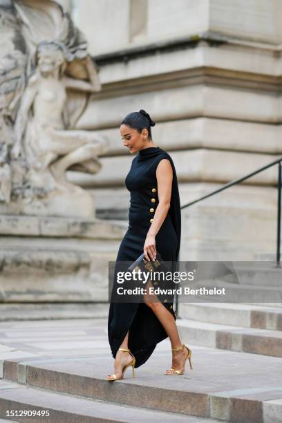 Amina Muaddi wears gold small earrings, a black wrap high neck / sleeveless / large gold buttons side long dress from Schiaparelli, gold large...