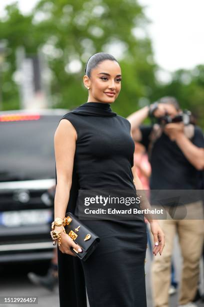 Amina Muaddi wears gold small earrings, a black wrap high neck / sleeveless / large gold buttons side long dress from Schiaparelli, gold large...