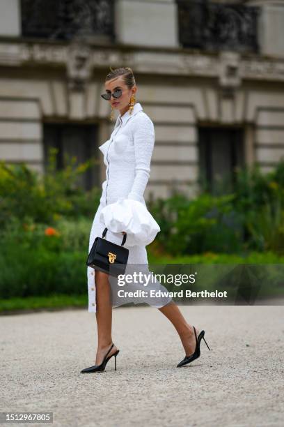 Xenia Adonts wears black with embroidered rhinestones cat eyes sunglasses, gold large ear pendant earrings from Schiaparelli, a white ruffled...