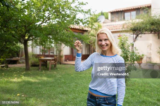 smiling mature woman holding key in front of house - clé de maison photos et images de collection