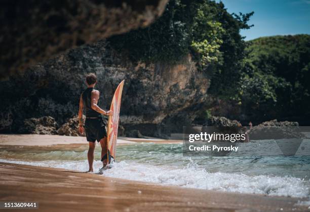 a male surfer walks along the sea with a surfboard - uluwatu stock pictures, royalty-free photos & images