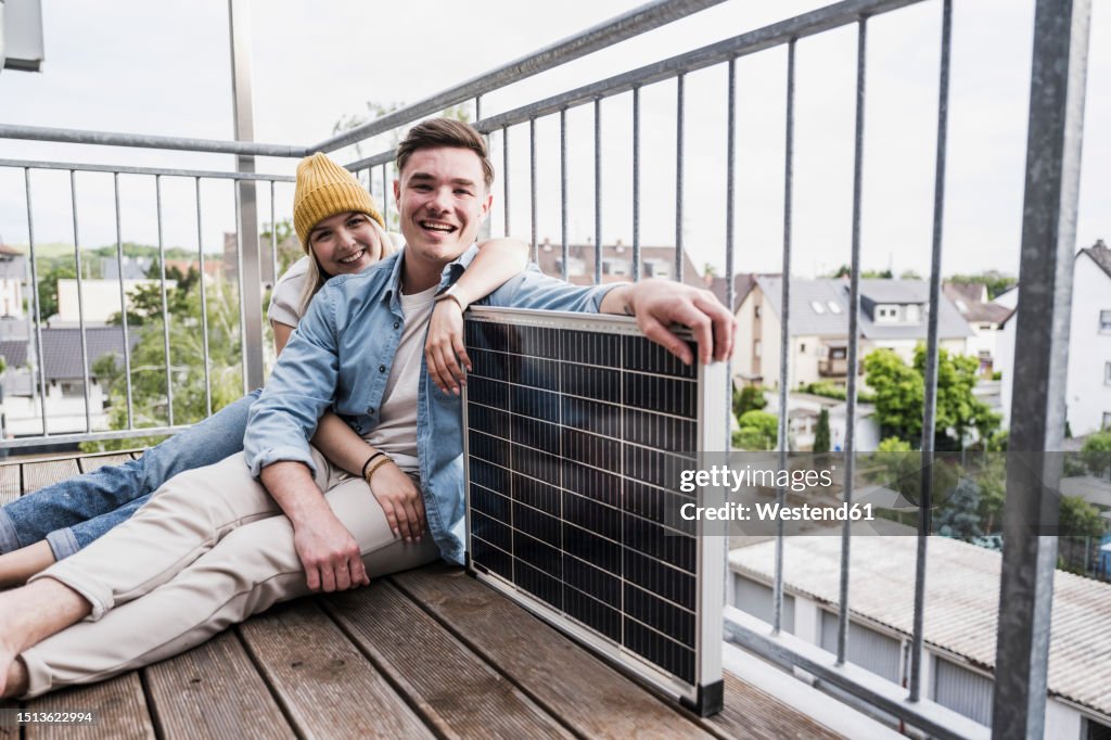 Happy young couple sitting with solar panel by railing on balcony