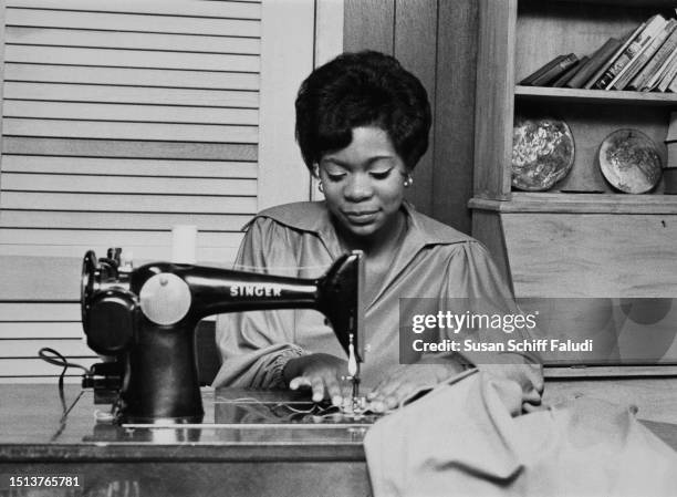 Woman sitting at a Singer sewing machine, sewing a length of fabric, with books and plates on the shelves behind her, United States, circa 1970.