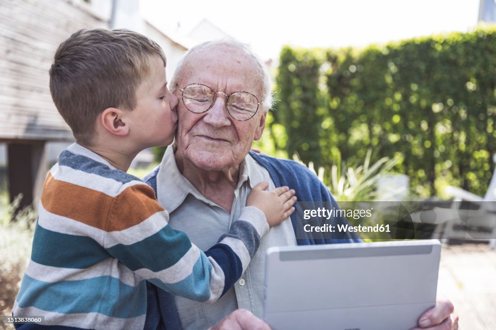 Boy kissing and embracing grandfather holding tablet PC