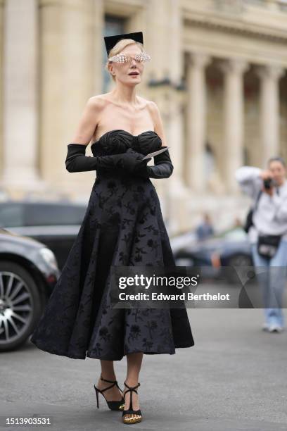 Guest wears a black velvet large squared headband, rhinestones glasses from Schiaparelli, a black silk with embroidered velvet pattern shoulder-off /...