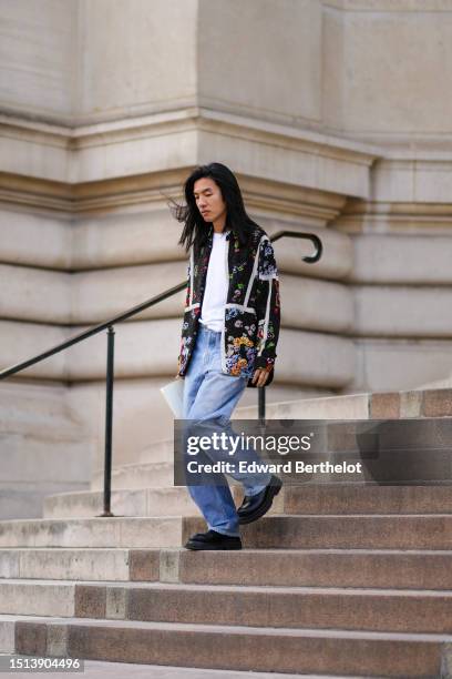 Guest wears a white t-shirt, a black with embroidered blue / green / red embroidered flower pattern jacket, blue faded denim large pants, black shiny...