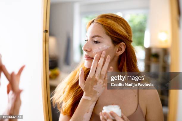 woman with vitiligo and acne on her face, doing a skin care while observing herself in the mirror - vitiligo stock pictures, royalty-free photos & images