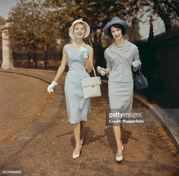 Exterior scene of two female fashion models wearing, from left, a pale blue sleeveless fitted dress with button decoration and a pale blue grey...