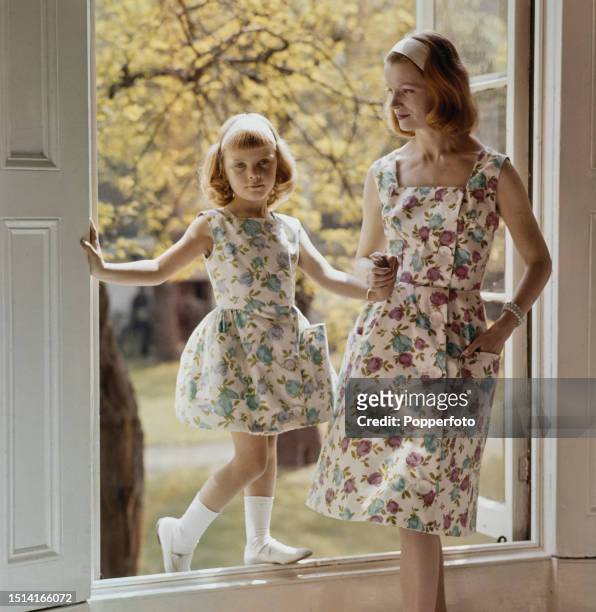 Female fashion model posed with a young girl beside an open window, they both wear matching button through summer dresses with a floral rose pattern,...