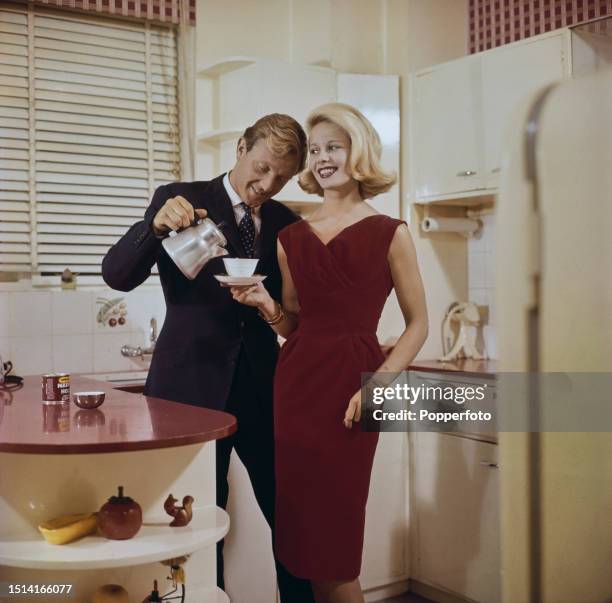 Female fashion model posed wearing a knee length red sleeveless velvet dress in a kitchen setting with a man, dressed in a dark suit, pouring coffee...