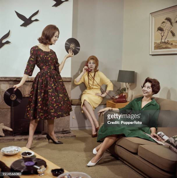 Three female fashion models posed in a living room setting, they wear winter dresses in red and brown pattern, pale yellow and dark green velvet,...