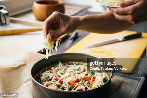 female baker adding grounded cheese on top of uncooked pie stuffed with tuna, broccoli and bell pepper lying in round form on baking sheet before baking. - espolvorear fotografías e imágenes de stock