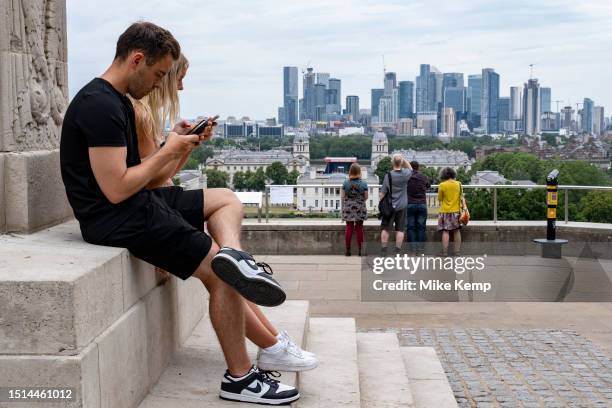 As tourists and visitors look out towards the skyline of Canary Wharf financial district from the viewpoint in Greenwich Park, two people sit...