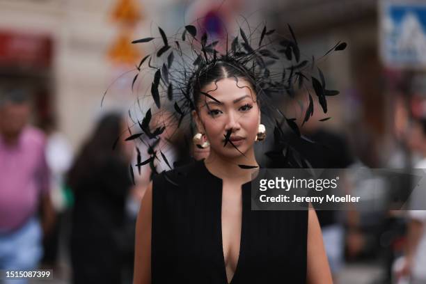 Fashion Week guest is seen wearing large golden hoop earrings, a headdress with black feathers standing out from her head, a black loose sleeveless...