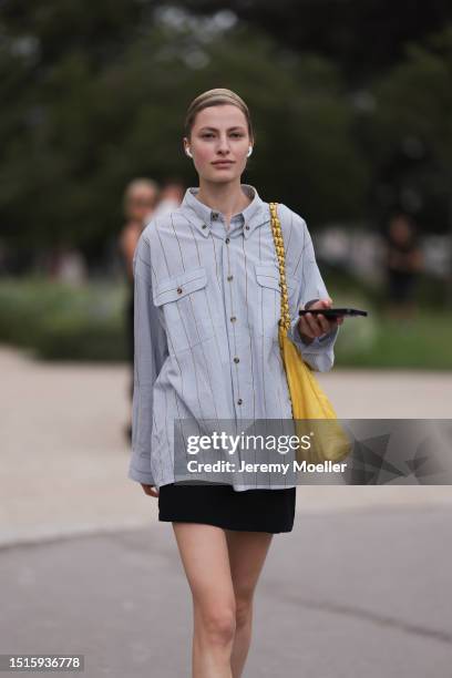 Guest seen outside Schiaparelli show wearing Apple Air Pod Pro, oversized blue shirt with stripes, yellow Chanel tote bag and black Adidas Gazelle...