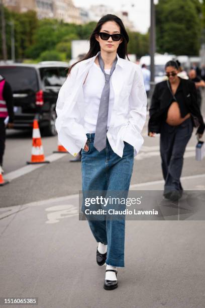 Model wears a white shirt, white tank top, grey Chanel tie, a long denim skirt and black mary jane shoes with white socks, outside Chanel, during the...