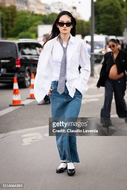 Model wears a white shirt, white tank top, grey Chanel tie, a long denim skirt and black mary jane shoes with white socks, outside Chanel, during the...