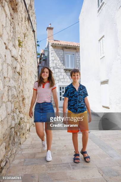 teenage girl and preadolescent boy walking in the stone paved alley on a hot summer day - boy skirt stock pictures, royalty-free photos & images