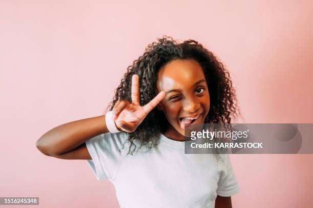 beautiful little afro girl sticking out her tongue and putting her fingers in a v shape on pink background. - kid putting finger in mouth stock-fotos und bilder