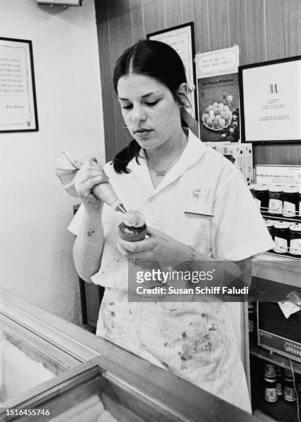 Young woman applies a whipped cream crown to an ice cream cone as she stands behind the counter of an ice cream parlour, United States, circa 1975.