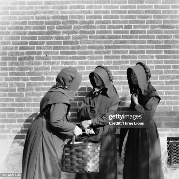 Three Amish women, one holding a basket, in matching outfits, comprising black full-length dresses, black aprons and black bonnets, with a brick wall...