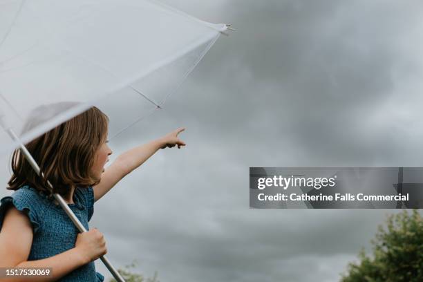 a child stands under a transparent umbrella, facing away from the camera. she points ip towards a heavy, dark sky. a rainstorm is about to begin. - tormenta eléctrica fotografías e imágenes de stock