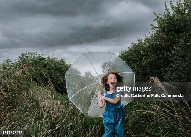 a little girl squeals happily and runs towards the viewer, clutching an umbrella, as she is caught in a heavy rainstorm - rainy season stock pictures, royalty-free photos & images