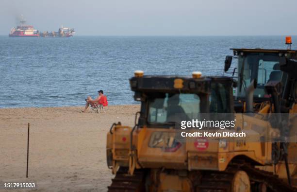 Person sits on the beach next to heavy machinery as the Army Corps of Engineers continues their beach and dune replenishment project on July 05, 2023...