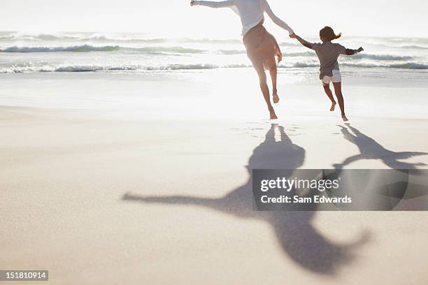 mother and daughter holding hands and running on sunny beach - running shadow stock-fotos und bilder