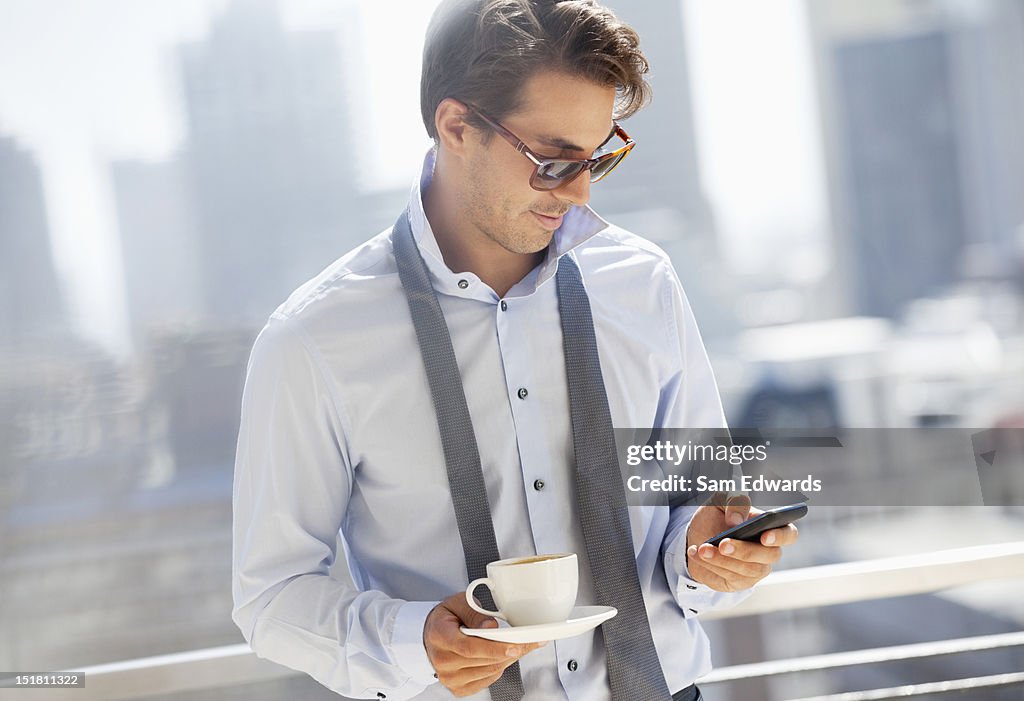 Businessman with coffee checking cell phone on urban balcony