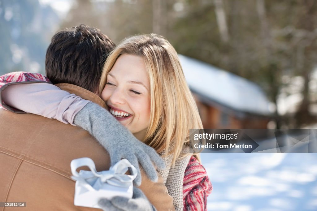 Smiling woman holding Christmas gift and hugging man in snowy field