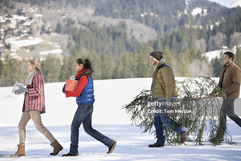 Couples and dog carrying fresh cut Christmas tree and gifts in snow
