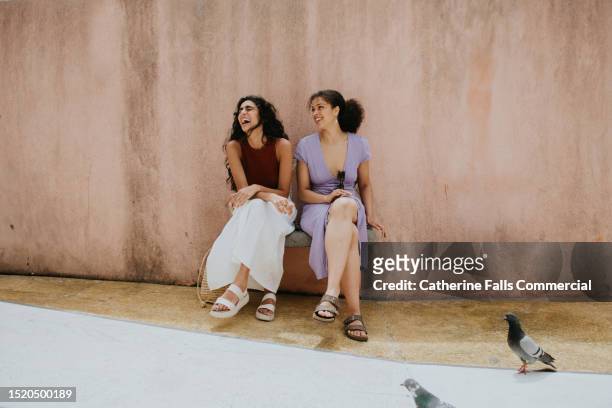 two woman sit on a bench against a wall and laugh - annual companions stock pictures, royalty-free photos & images