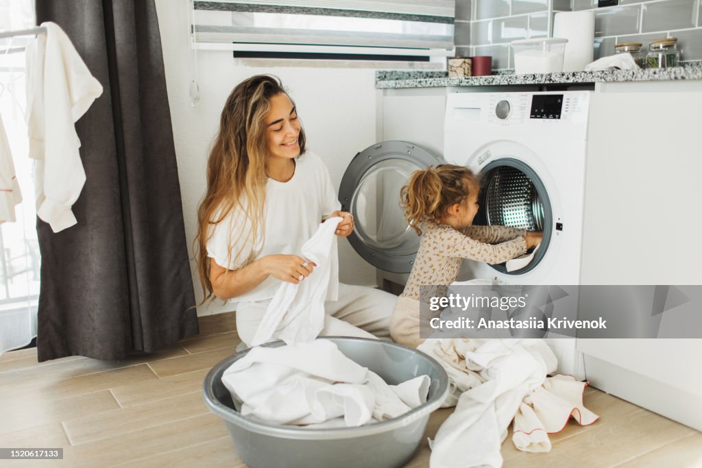 Mother a housewife with a baby engaged in laundry fold clothes into the washing machine.