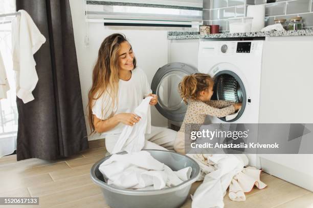 mother a housewife with a baby engaged in laundry fold clothes into the washing machine. - white laundry foto e immagini stock