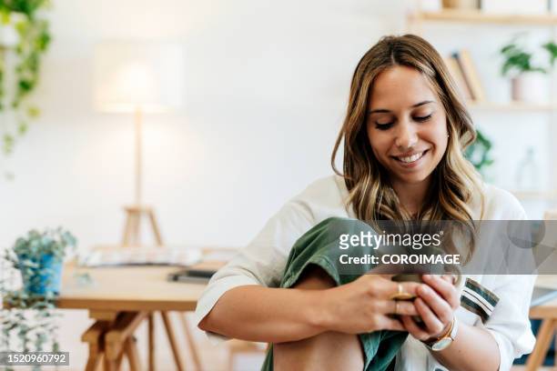 woman sitting relaxed in a chair in her home office drinking a cup of coffee and looking down. 
woman happy with herself. - leisure work coffee happy stockfoto's en -beelden