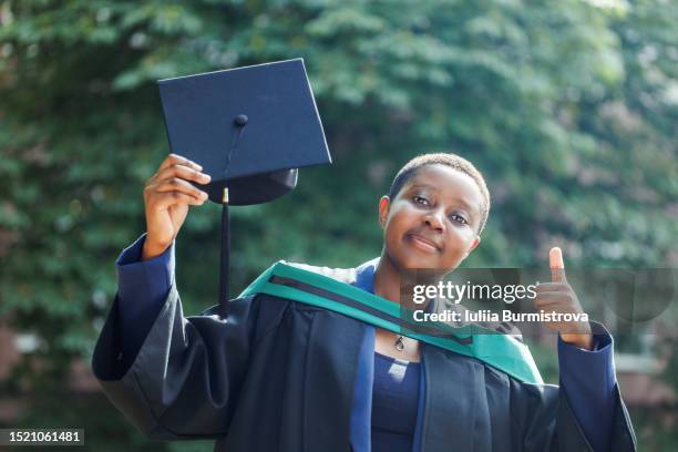 positive young black lady with short black hair in academic regalia standing and holding diploma giving thumbs up - scholarship award stock pictures, royalty-free photos & images