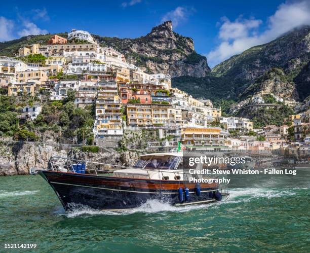 classic motorboat passes the scenic town of positano set amongst the beautiful hills of the amalfi coast in italy - costiera amalfitana foto e immagini stock