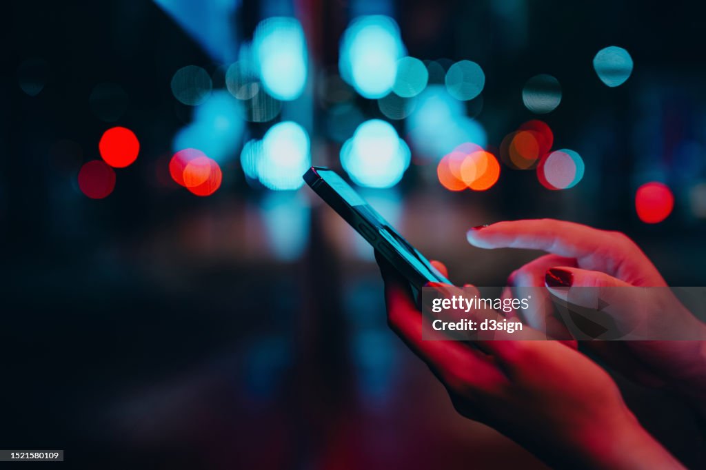Young woman networking with social media on smartphone in city street at night. Defocus street lights in background. Close up of woman checking social media on smartphone in the city. People engaging in networking with technology. Lifestyle and technology