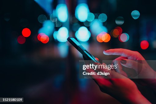 Young woman networking with social media on smartphone in city street at night. Defocus street lights in background. Close up of woman checking social media on smartphone in the city. People engaging in networking with technology. Lifestyle and technology Young woman networking with social media on smartphone in city street at night. Defocus street lights in background. Close up of woman checking social media on smartphone in the city. People engaging in networking with technology. Lifestyle and technology