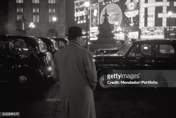 Man wearing a bowler in Piccadilly Circus, in the traffic of cars and lights. London , 1957.